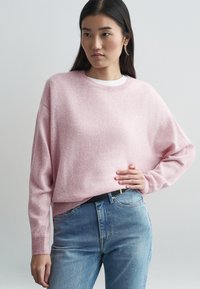 Pulover tricotat roz deschis, cu un croi lejer, guler rotund și mâneci lungi. Asortat cu blugi albaștri cu talie înaltă și o curea neagră.