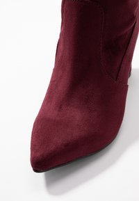 Botín de ante burdeos con puntera puntiaguda, con textura suave y diseño minimalista, adecuado para diversas ocasiones.