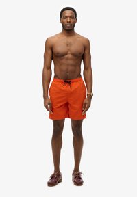 Bañador naranja con cordón negro ajustable, bolsillos laterales y un sutil patrón texturizado. Llevado con zapatos marrones con cordones.