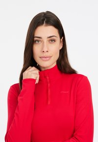 Červený top s dlhým rukávom a polo-vystrihom, obsahujúci polozips a detail loga. Hladká tkanina s priliehavým strihom a jednotnou farbou.