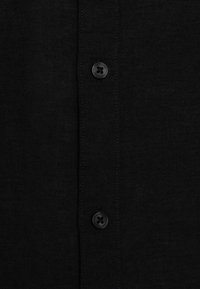 Schwarzes Hemd aus strukturiertem Stoff, mit Knopfleiste und einem runden, schwarzen Knopf mit eingraviertem Logo. Einfaches und minimalistisches Design.