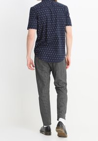 Camicia navy a maniche corte con un motivo geometrico bianco, abbinata a pantaloni grigi a quadretti e scarpe nere, vista da dietro.
