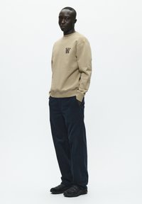 Beige sweatshirt med et lille sort logo på brystet, parret med mørkeblå kordfløjspants og sorte strukturerede sko. Model står foran en hvid baggrund.