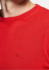 Pull en laine rouge avec un col rond, arborant un logo subtil brodé sur la poitrine. La texture est lisse avec des coutures serrées.