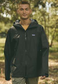 Czarna wodoodporna kurtka z zamkiem błyskawicznym z przodu, kapturem i logo Patagonia na piersi. Model ma na sobie żółtą koszulę pod spodem.