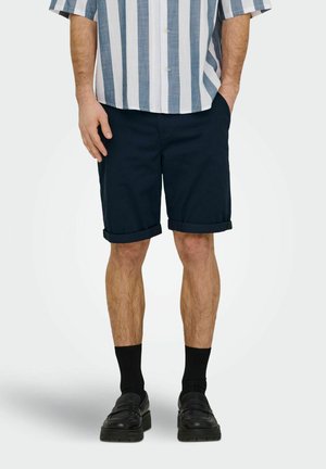 Férfi, aki felhajtott sötétkék rövidnadrágot, csíkos rövid ujjú gombos inget, fekete zoknit és fekete mokaszint visel, és zsebében tartja a kezét.