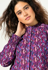 Blusa morada con un patrón ondulado en rosa, negro y azul. Presenta un escote y puños fruncidos, tejido texturizado y cierre de botones en la parte delantera.