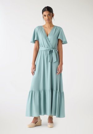 Donna in piedi che indossa un vestito avvolgente azzurro chiaro con maniche svolazzanti e cintura annodata in vita, abbinato a espadrillas beige con zeppa.