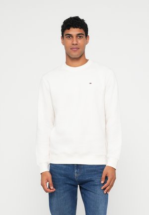 Sweatshirt crème clair à manches longues, col rond, poignets et ourlet côtelés. Présente un petit logo sur la poitrine ; porté avec un jean bleu.