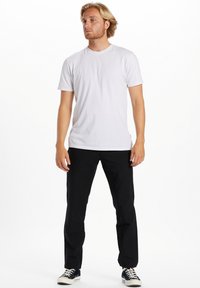 Hvid bomulds T-shirt med rund hals, parret med sorte bukser. Modellen står med en neutral kropsholdning og navy sko med hvide detaljer.