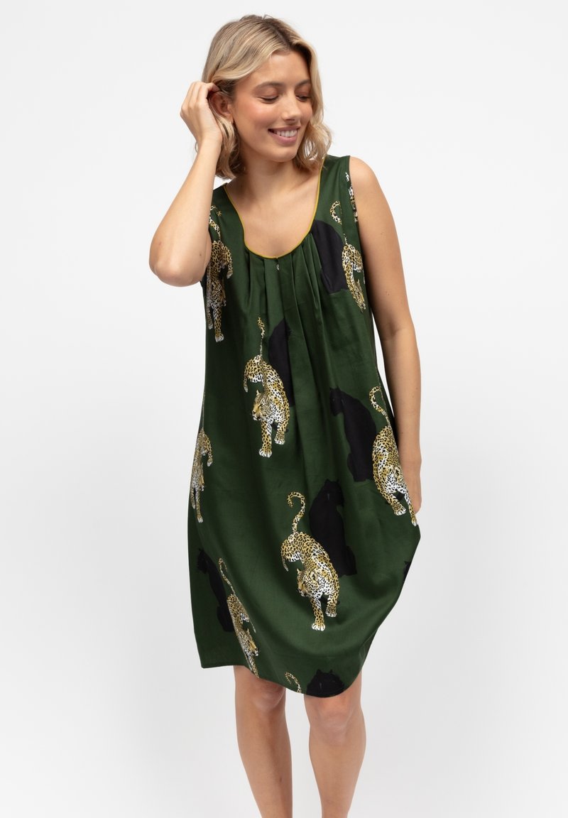 Donna bionda che sorride e si tocca i capelli, indossando un vestito senza maniche di colore verde scuro con un motivo maculato nero e giallo.