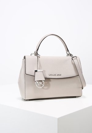 Jasnobeżowa torebka Michael Kors z górnym uchwytem, klapkowym zamknięciem, odpinanym paskiem na ramię i zawieszką z logo MK, umieszczona na białym postumencie.