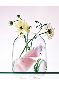 Roze cosmeticafles met gouden tekst, gedeeltelijk ondergedompeld in water, onder een glazen koepel. Omringd door groene stelen en lichtgele bloemen.