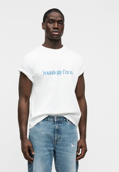 Άνδρας που στέκεται μπροστά σε απλό φόντο, φοράει λευκό μπλουζάκι με γυρισμένα μανίκια και μπλε κείμενο «Sounds gay I'm in», συνδυασμένο με ανοιχτόχρωμο μπλε τζιν.