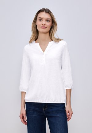 Jeune femme portant un t-shirt Henley blanc à manches trois-quarts et un jean bleu foncé, debout devant un fond gris neutre.