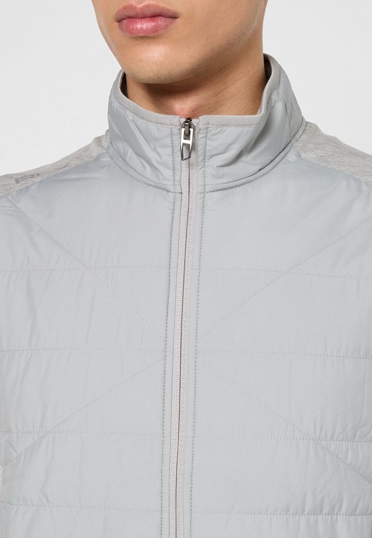 Chaqueta acolchada de un gris claro con un cuello alto, que cuenta con un cierre delantero y acentos en los hombros de un gris contrastante. Tejido suave y ligero.