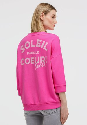 Blonde Frau trägt einen leuchtend pinken Sweatshirt mit dem Text "SOLEIL DANS LE COEUR! soccx" auf dem Rücken, kombiniert mit hellgrauen Jeans.