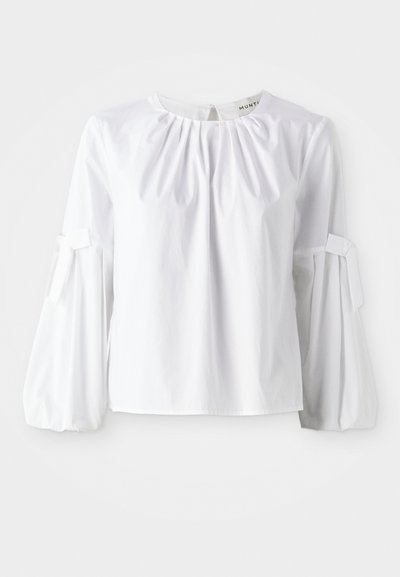 Fehér, hosszú ujjú pamut felső, kerek nyakkivágással, diszkrét redőkkel és megkötős mandzsettákkal, amelyek strukturált formát és tekstúrát adnak.