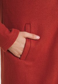 Rode wollen jas met een zachte textuur, voorzien van een zijzak en een aansluitende manchette. De stof lijkt warm en gezellig, ideaal voor koudere weersomstandigheden.