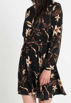 Kvinde iført en sort langærmet kjole med blomsterprint og et bindebælte i taljen, stående mod en ensfarvet lys baggrund.
