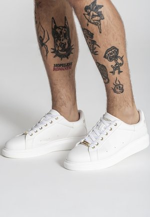 Piernas con varios tatuajes negros, que incluyen la cabeza de un perro, una araña, un ángel y el texto "Romántico Desesperado", llevando zapatillas blancas con cordones y detalles dorados.