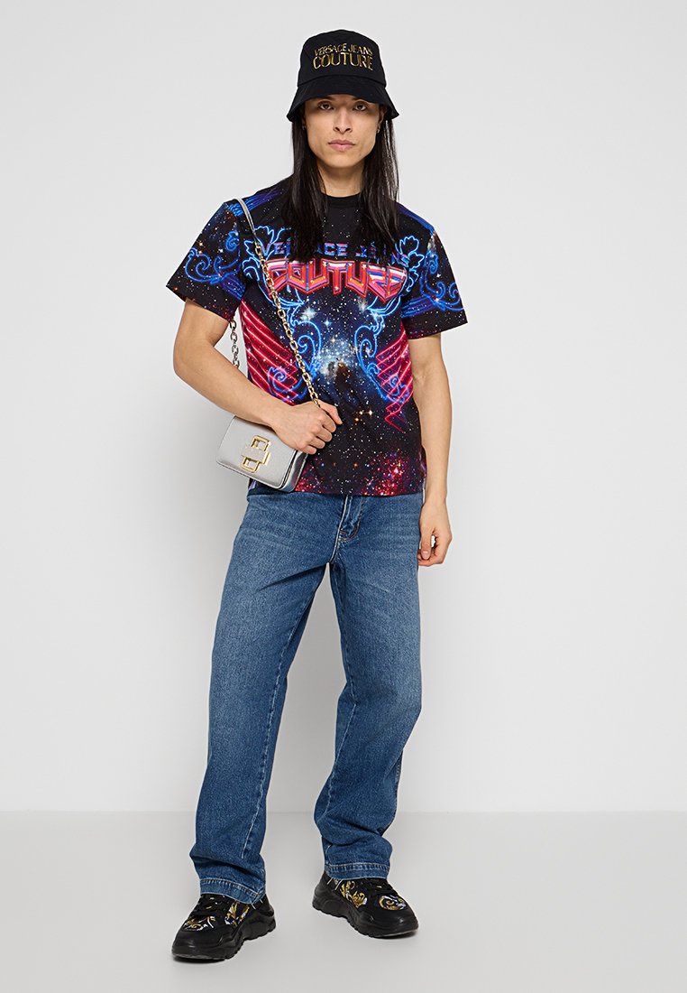 Camiseta negra con estampado cósmico multicolor y texto "COUTURE", gorra tipo bucket azul y negra, jeans azules y zapatillas negras con un patrón y un pequeño bolso.