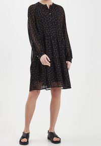 Černé šaty s puntíky z průhledné látky; dlouhé rukávy, kulatý výstřih, vrstvený design. Nosí se s černými sandály, což vytváří uvolněný styl.