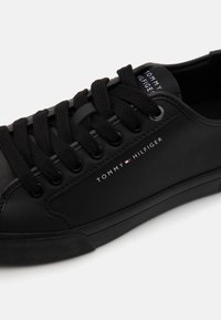 Zapatilla de cuero negro con punta redonda, suela plana de goma, cordones negros y el logo de Tommy Hilfiger impreso en blanco. Diseño minimalista.