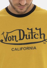 Homme portant un t-shirt jaune moutarde avec un logo noir "Von Dutch California" et un col noir, vu de la nuque à la poitrine.