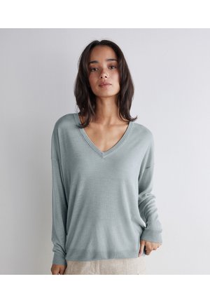 Kvinde med skulderlangt mørkt hår iført en lyseblå V-hals sweater og beige bukser mod en ensfarvet hvid baggrund.