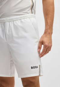 Pantalones cortos deportivos blancos con cintura elástica, bolsillos laterales y un logo negro de "BOSS". Presenta rayas verticales en contraste a los lados.