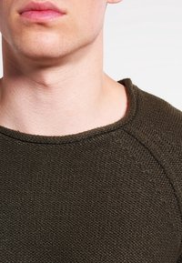 Sweat-shirt en maille vert olive foncé avec une surface texturée, doté d'un col rond et de détails de coutures sur les épaules.