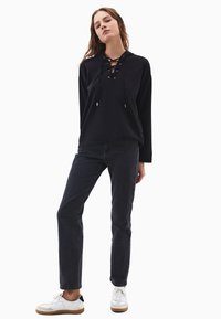 Černá mikina s kapucí a šněrovacím výstřihem, kombinovaná s tmavými džíny a bílými teniskami. Materiál vypadá měkce a má uvolněný střih.