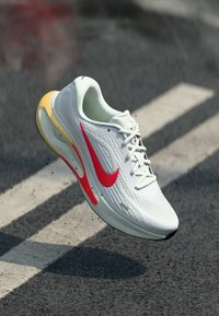 Λευκό αθλητικό παπούτσι με κόκκινο λογότυπο Nike, με υφασμένο επάνω μέρος, επενδυμένο γιακά και χαρακτηριστική κίτρινη σόλα.