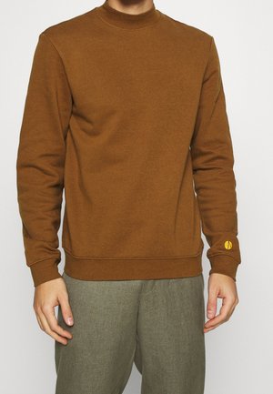 Mann trägt braunes Sweatshirt mit langen Ärmeln und grau-grüne Hose, steht mit entspannten Armen an den Seiten vor einem schlichten Hintergrund.