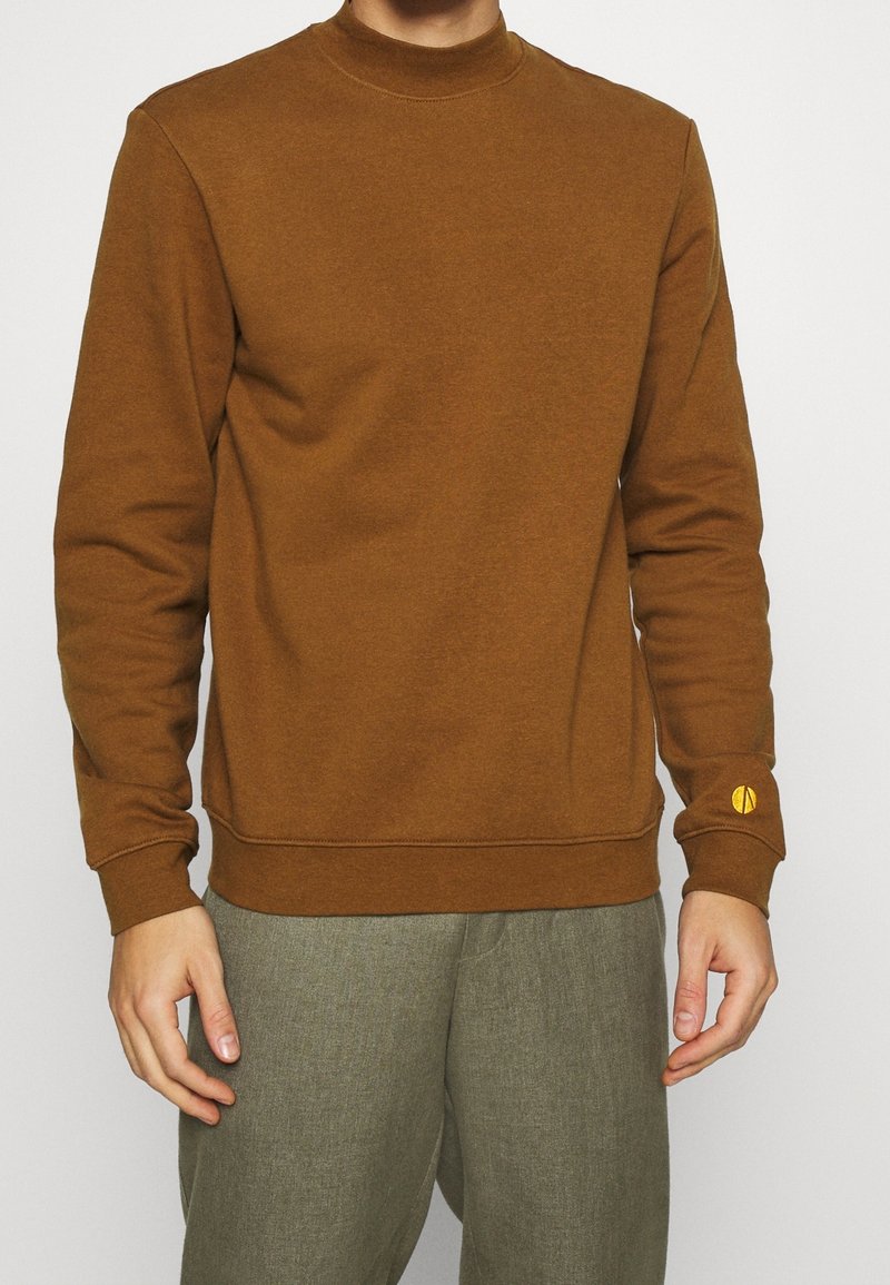 Homme portant un sweat-shirt marron à manches longues et un pantalon gris-vert, debout avec les bras détendus le long du corps sur un fond uni.