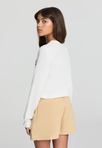 Top bianco a maniche lunghe con scollo rotondo, abbinato a pantaloni corti beige. Presenta una sottile trama e una cintura elastica sui pantaloni.
