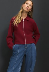Pull à zip bordeaux avec col et manches longues, associé à un jean en denim bleu taille haute. Tissu lisse, design simple.