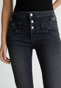 Jeans negros de tiro alto con un diseño ajustado, que presentan un cierre de tres botones y detalles metálicos en la cinturilla. Textura de denim suave.