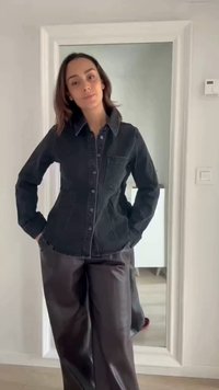 Femme aux cheveux mi-longs porte une veste en jean foncée et un pantalon large en cuir, debout les mains dans les poches devant un miroir.