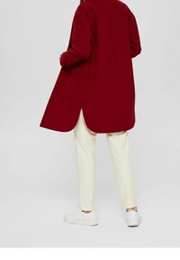 Długi, czerwony płaszcz o prostym kroju z bocznymi kieszeniami, zestawiony z kremowymi spodniami i białymi trampkami, na neutralnym tle.