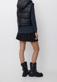 Doudoune sans manches noire avec capuche, haut à manches longues bleu marine, jupe plissée noire, et bottes en cuir noires montantes jusqu'aux genoux avec boucles et semelles texturées.