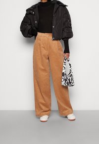 Personen bär svart kort pufferjacka, svart turtleneck, vida beige kordbyxor, vita sneakers och håller en svart-vit mönstrad handväska.