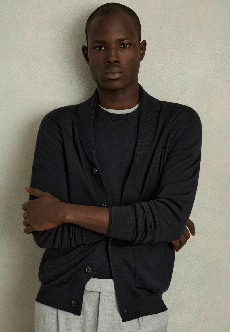 Cardigan gris oscuro con cuello de chal, cierres de botón y puños acanalados, llevado sobre una camisa gris, combinado con pantalones gris claro.