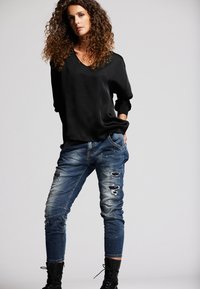 Schwarzes, lockeres Langarmoberteil mit V-Ausschnitt. Zerrissene blaue Jeans aus Denim mit Rissen, schmal zulaufendem Bein und schwarzen Schnürstiefeln.