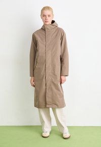 Ljusbrun vattentät jacka med hög krage och huva, med långa ärmar och sidofickor, bärs över lösa beige byxor.