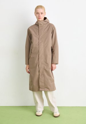 Lichtbruine waterdichte jas met hoge kraag en capuchon, met lange mouwen en zijzakken, gedragen over losse beige broek.