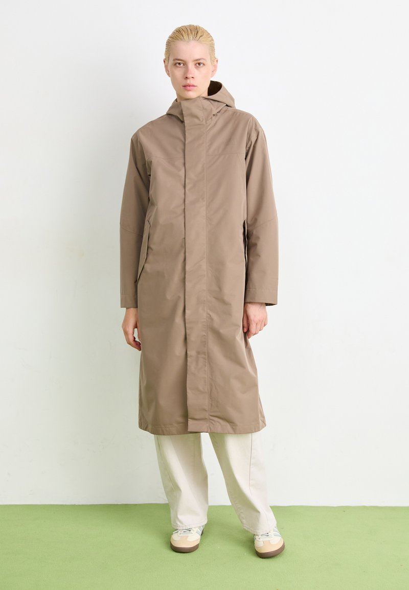 Ljusbrun vattentät jacka med hög krage och huva, med långa ärmar och sidofickor, bärs över lösa beige byxor.