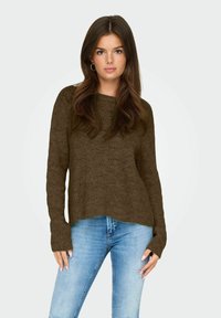 Jeune femme avec de longs cheveux bruns portant des boucles d'oreilles créoles, un pull en tricot marron avec des ouvertures pour les pouces, et un jean bleu clair déchiré.