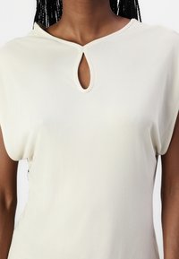 Top beige clair avec un décolleté en trou de serrure, des manches courtes et un tissu lisse et texturé. Présente une coupe décontractée et un design minimaliste.